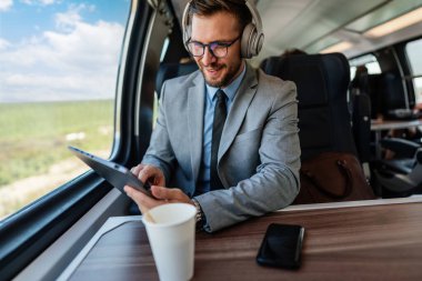 Genç işadamı kulaklık takıyor ve trenle seyahat ederken dijital tablet kullanıyor. Müzikten zevk alıyor ve iş seyahati sırasında üretken kalıyor.