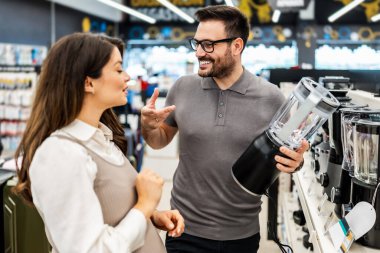 Neşeli bir elektronik mağazasında çeşitli ürünler sergilerken hamile bir müşteriye ev aletleri seçmesinde yardımcı olan gülümseyen mağaza asistanı