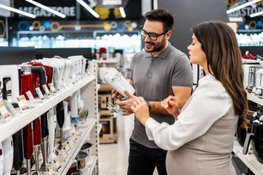 Neşeli bir elektronik mağazasında çeşitli ürünler sergilerken hamile bir müşteriye ev aletleri seçmesinde yardımcı olan gülümseyen mağaza asistanı