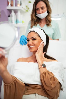 Güzel ve mutlu genç bir kadın tıbbi sandalyede oturup aynaya bakıyor. Hyaluronik asit doldurucu ya da botulinyum toksin iğneleriyle başarılı bir güzellik tedavisinden sonra tatmin oluyor..