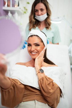 Güzel ve mutlu genç bir kadın tıbbi sandalyede oturup aynaya bakıyor. Hyaluronik asit doldurucu ya da botulinyum toksin iğneleriyle başarılı bir güzellik tedavisinden sonra tatmin oluyor..