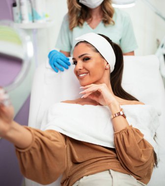 Güzel ve mutlu genç bir kadın tıbbi sandalyede oturup aynaya bakıyor. Hyaluronik asit doldurucu ya da botulinyum toksin iğneleriyle başarılı bir güzellik tedavisinden sonra tatmin oluyor..