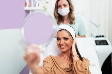 Güzel ve mutlu genç bir kadın tıbbi sandalyede oturup aynaya bakıyor. Hyaluronik asit doldurucu ya da botulinyum toksin iğneleriyle başarılı bir güzellik tedavisinden sonra tatmin oluyor..