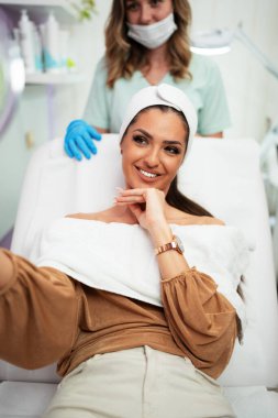 Güzel ve mutlu genç bir kadın tıbbi sandalyede oturup aynaya bakıyor. Hyaluronik asit doldurucu ya da botulinyum toksin iğneleriyle başarılı bir güzellik tedavisinden sonra tatmin oluyor..