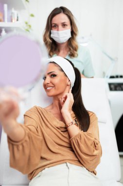 Güzel ve mutlu genç bir kadın tıbbi sandalyede oturup aynaya bakıyor. Hyaluronik asit doldurucu ya da botulinyum toksin iğneleriyle başarılı bir güzellik tedavisinden sonra tatmin oluyor..