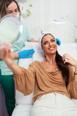 Güzel ve mutlu genç bir kadın tıbbi sandalyede oturup aynaya bakıyor. Hyaluronik asit doldurucu ya da botulinyum toksin iğneleriyle başarılı bir güzellik tedavisinden sonra tatmin oluyor..