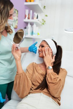 Mavi eldiven takan bir güzellik uzmanı, bir güzellik salonundaki yüz tedavisinin sonuçlarını belirtirken, kadın da aynada mutlu ve memnun görünüyor.