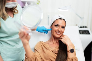 Mavi eldiven takan bir güzellik uzmanı, bir güzellik salonundaki yüz tedavisinin sonuçlarını belirtirken, kadın da aynada mutlu ve memnun görünüyor.