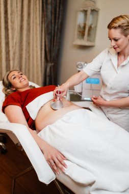 Güzellik uzmanı, profesyonel güzellik merkezinde yatan gülümseyen olgun bir kadının karnına radyasyon kaldırma cihazı uyguluyor. Modern, zararsız bir vücut şekillendirme tedavisi sunuyor..