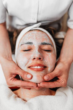 Profesyonel güzellik uzmanı gevşemiş bir müşterinin yüzüne nazikçe kremsi bir maske takarak sağlıklı cilt için rahatlatıcı ve canlandırıcı bir spa tedavisi uyguluyor.