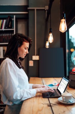 Güzel bir bayan öğrenci ya da genç bir iş kadını dizüstü bilgisayarını kullanıyor ve rahat bir kafede pencerenin yanında duruyor..