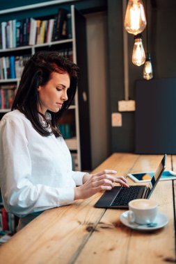 Güzel bir bayan öğrenci ya da genç bir iş kadını dizüstü bilgisayarını kullanıyor ve rahat bir kafede pencerenin yanında duruyor..