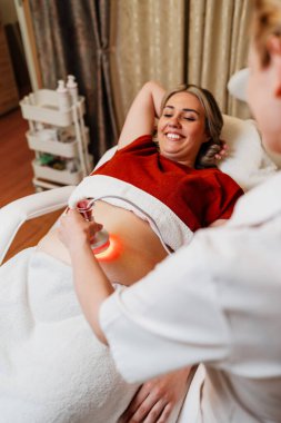 Modern güzellik merkezinde müşterinin karnına kırmızı ışık tedavisi uygulayan profesyonel güzellik uzmanı cilt gençleştirme ve kolajen üretimini teşvik ediyor.