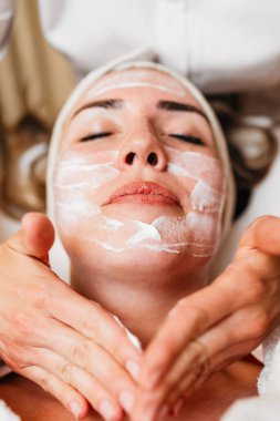 Profesyonel güzellik uzmanı gevşemiş bir müşterinin yüzüne nazikçe kremsi bir maske takarak sağlıklı cilt için rahatlatıcı ve canlandırıcı bir spa tedavisi uyguluyor.