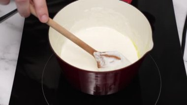 Şef, indüksiyon sobasının üstünde kırmızı bir tencerede, ahşap bir kaşıkla kremalı sosu çırpıyor ve leziz bir mutfak yaratımı için pürüzsüz ve lezzetli bir taban oluşturuyor.