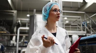 Koruyucu giysiler ve maske takan kadın kalite kontrol müfettişi su şişeleme fabrikasındaki üretim hattını incelerken panoya notlar yazıyor.