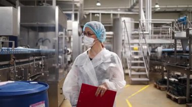 Koruyucu giysiler ve maske takan kadın kalite kontrol müfettişi su şişeleme fabrikasındaki üretim hattını incelerken panoya notlar yazıyor.