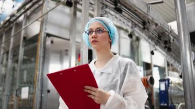 Koruyucu giysiler ve maske takan kadın kalite kontrol müfettişi su şişeleme fabrikasındaki üretim hattını incelerken panoya notlar yazıyor.