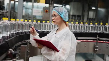 Koruyucu giysiler ve maske takan kadın kalite kontrol müfettişi su şişeleme fabrikasındaki üretim hattını incelerken panoya notlar yazıyor.