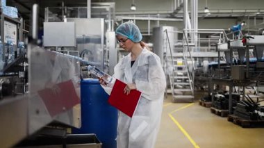 Koruyucu giysiler ve maske takan kadın kalite kontrol müfettişi su şişeleme fabrikasındaki üretim hattını incelerken panoya notlar yazıyor.