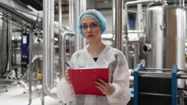 Koruyucu giysiler ve maske takan kadın kalite kontrol müfettişi su şişeleme fabrikasındaki üretim hattını incelerken panoya notlar yazıyor.