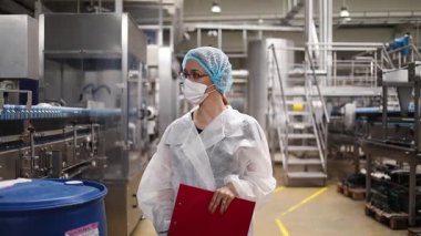Koruyucu giysiler ve maske takan kadın kalite kontrol müfettişi su şişeleme fabrikasındaki üretim hattını incelerken panoya notlar yazıyor.