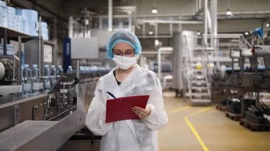 Koruyucu giysiler ve maske takan kadın kalite kontrol müfettişi su şişeleme fabrikasındaki üretim hattını incelerken panoya notlar yazıyor.