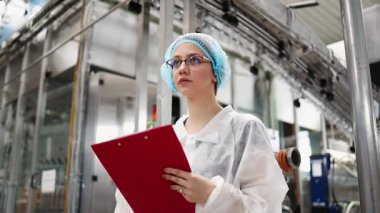 Koruyucu giysiler ve maske takan kadın kalite kontrol müfettişi su şişeleme fabrikasındaki üretim hattını incelerken panoya notlar yazıyor.