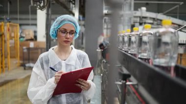Koruyucu giysiler ve maske takan kadın kalite kontrol müfettişi su şişeleme fabrikasındaki üretim hattını incelerken panoya notlar yazıyor.