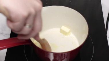 Şef, indüksiyon sobasının üstünde kırmızı bir tencerede, ahşap bir kaşıkla kremalı sosu çırpıyor ve leziz bir mutfak yaratımı için pürüzsüz ve lezzetli bir taban oluşturuyor.