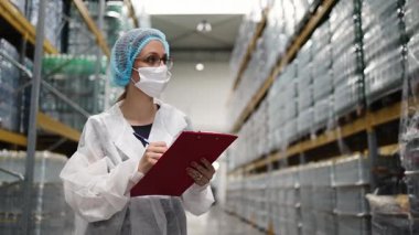 Dağıtım deposundaki raflarda depolanan ürün ve eşyaları incelerken, laboratuvar önlüğü ve saç filesi ile panoya notlar yazan kadın denetçi.