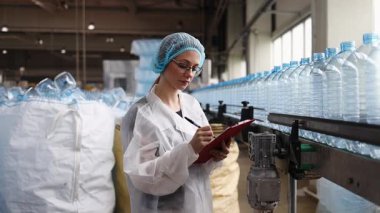 Koruyucu giysiler ve maske takan kadın kalite kontrol müfettişi su şişeleme fabrikasındaki üretim hattını incelerken panoya notlar yazıyor.