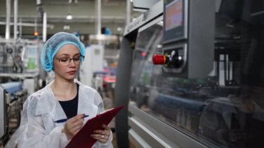 Koruyucu giysiler ve maske takan kadın kalite kontrol müfettişi su şişeleme fabrikasındaki üretim hattını incelerken panoya notlar yazıyor.