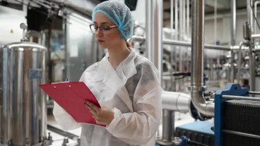 Koruyucu giysiler ve maske takan kadın kalite kontrol müfettişi su şişeleme fabrikasındaki üretim hattını incelerken panoya notlar yazıyor.