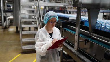 Koruyucu giysiler ve maske takan kadın kalite kontrol müfettişi su şişeleme fabrikasındaki üretim hattını incelerken panoya notlar yazıyor.