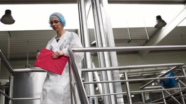 Koruyucu iş kıyafetlerinde çalışan, tıbbi malzeme araştırma ve üretim fabrikasında çalışan kadın işçi. Kontrol köprüsünde duruyor veya yürüyor ve üretim hattını izliyor..