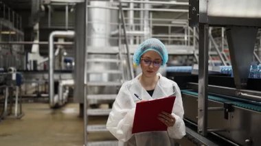 Koruyucu giysiler ve maske takan kadın kalite kontrol müfettişi su şişeleme fabrikasındaki üretim hattını incelerken panoya notlar yazıyor.