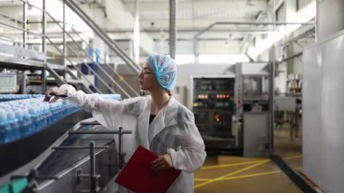 Koruyucu giysiler ve maske takan kadın kalite kontrol müfettişi su şişeleme fabrikasındaki üretim hattını incelerken panoya notlar yazıyor.