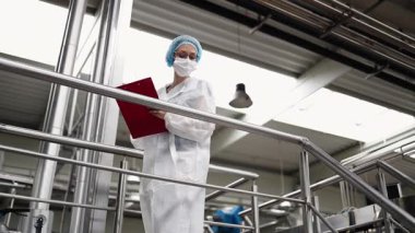 Koruyucu iş kıyafetlerinde çalışan, tıbbi malzeme araştırma ve üretim fabrikasında çalışan kadın işçi. Kontrol köprüsünde duruyor veya yürüyor ve üretim hattını izliyor..