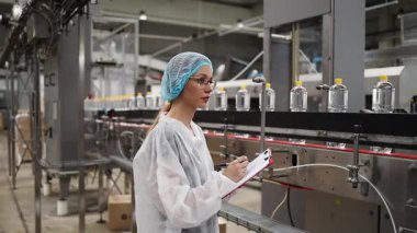 Koruyucu giysiler ve maske takan kadın kalite kontrol müfettişi su şişeleme fabrikasındaki üretim hattını incelerken panoya notlar yazıyor.