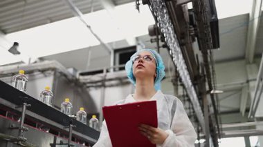 Koruyucu giysiler ve maske takan kadın kalite kontrol müfettişi su şişeleme fabrikasındaki üretim hattını incelerken panoya notlar yazıyor.