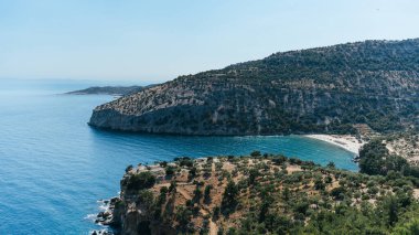 Dik kayalıkları saran yemyeşil bitki örtüsü, Yunanistan 'ın Thasos adasındaki gözlerden uzak bir kumsala nazikçe uzanan kristal berraklığında turkuaz suyla çevrili bir körfezi kaplıyor.