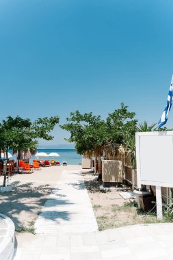 Güneşli yataklar, şemsiyeler, saman plaj kulübeleri, rüzgarda dalgalanan bir Yunan bayrağı ve açık mavi gökyüzünün altında yemyeşil ağaçlar olan kumlu bir sahile giden beyaz ahşap bir yol. Thasos adası. Yunanistan.