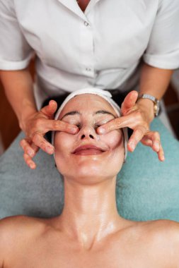 Profesyonel güzellik uzmanı masaj masasına uzanmış kadın müşterinin yüzüne nazikçe masaj yapıyor. Gözleri kapalı, güzellik salonunda rahatlatıcı tedavinin keyfini çıkarıyor.