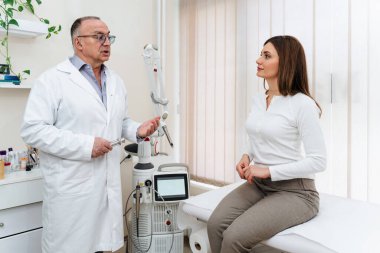 Cildiyeci modern bir klinikte muayene masasında oturan kadın hastaya lazer tedavi prosedürünü anlatıyor, kozmetik prosedürlerinin seçenekleri ve faydalarını tartışıyor.