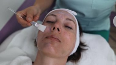 Profesyonel güzellik uzmanı gevşemiş bir müşterinin yüzüne nazikçe kremsi bir maske takarak sağlıklı cilt için rahatlatıcı ve canlandırıcı bir spa tedavisi uyguluyor.