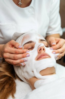 Güzellik uzmanı, güzellik salonunda gözleri kapalı yatan ve rahatlatıcı bir tedaviden zevk alan genç sarışın bir kadının yüzüne nazikçe köpük maske takıyor.