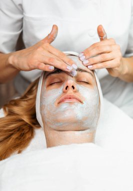 Güzellik uzmanı, tedavi maskesine uzanmış rahat bir kadına canlandırıcı yosun maskesi takarak profesyonel güzellik salonunda cilt bakımını arttırıyor.