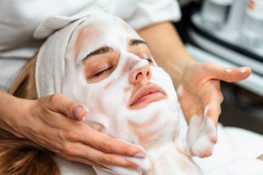 Güzellik uzmanı, güzellik salonunda gözleri kapalı yatan ve rahatlatıcı bir tedaviden zevk alan genç sarışın bir kadının yüzüne nazikçe köpük maske takıyor.