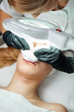 Siyah eldiven giyen bir güzellik uzmanı, yüz estetik ameliyatı sırasında ameliyat masasında kağıt mendille yatan genç bir kadının burnundaki sivilceyi çıkarıyor.